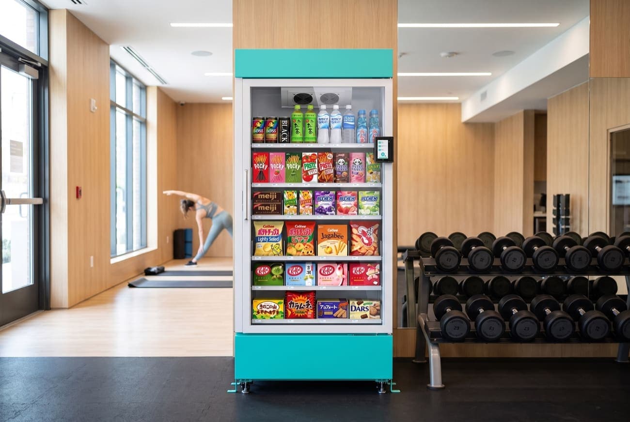 The Stocko fridge beside a dumbbell rack in a modern gym, someone stretching in the background